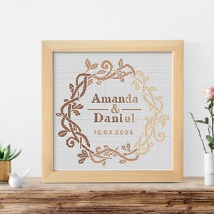 Op de afbeelding: Een ingelijst kruissteekkunstwerk met een kransontwerp in bruin- en goudtinten. De namen "Amanda & Daniel" en de datum "12.03.2025" staan in het midden van de krans. De lijst is gemaakt van lichtgekleurd hout.
