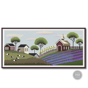May include: A framed cross-stitch landscape showing a rural scene with houses, a church, and a lavender field. Green trees and rolling hills are also featured. The artwork is in a dark brown frame.