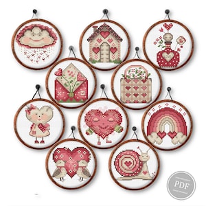 Könnte beinhalten: Zehn runde Kreuzstich-Designs mit Holzrahmen, jedes mit Valentinsthemen. Designs umfassen Herzen, ein Haus, eine Wolke, eine Schnecke und einen Regenbogen, alle in Rot-, Rosa- und Weißtönen. Die Designs sind alle gerahmt.