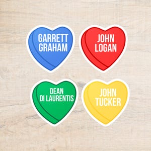 Könnte beinhalten: Vier herzförmige Aufkleber in Blau, Rot, Grün und Gelb. Jeder Aufkleber hat einen weißen Rand und einen Namen in weißem Text: Garrett Graham, John Logan, Dean Di Laurentis und John Tucker. Auf einer hellen Holzmaserung.
