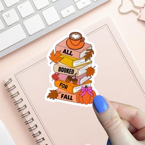 Peut inclure: Un autocollant représentant une pile de livres avec l'inscription "ALL BOOKED FOR FALL". Les livres sont ornés de feuilles d'automne, de glands et d'une citrouille. Une tasse de café avec un motif de cœur est posée dessus. L'autocollant est tenu par une main avec du vernis à ongles bleu.