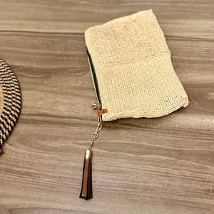 May include: A cream-colored woven clutch with a rectangular shape and a zipper closure. The clutch features a decorative tassel with brown and black leather strips. The bag is made of a textured material.