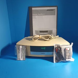 May include: A beige Bose Wave Radio II with a digital clock display showing 4:49, a power cord, a user manual, and two remote controls in plastic packaging. One remote has a label stating Battery Installed, and the other includes a black audio cable.