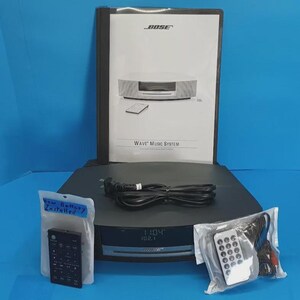 May include: A Bose Wave Music System with a black power cord, a user manual, and two remote controls in plastic bags. One bag has a label reading New Battery Installed. The system features a digital display showing 11:04 and 102.1.