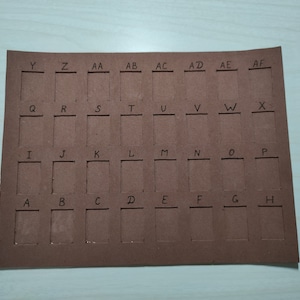May include: A brown cardboard sheet with cut-out squares labeled with letters from A to Z and then AA to AF. The squares are arranged in a grid pattern.