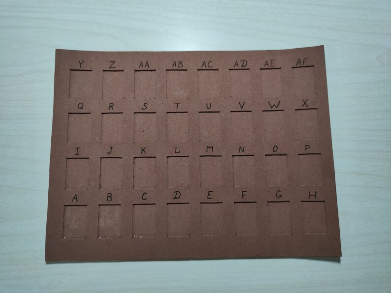 May include: A brown cardboard sheet with cut-out squares labeled with letters from A to Z and then AA to AF. The squares are arranged in a grid pattern.