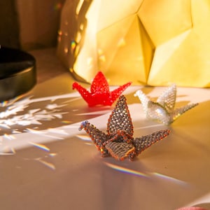 May include: Three origami paper cranes made of beads. One is red, one is white, and one is gray with orange accents.