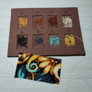 May include: A close-up of a beading board with various colors of beads in small containers labeled A through H. The board is brown and has a white thread attached to the container labeled D. A beaded pattern is visible below the board.