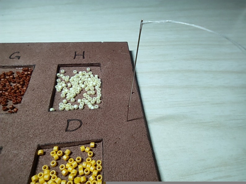 May include: A close-up of a brown board with different colored beads in separate compartments labeled with the letters G, H, and D. A needle and thread are visible in the upper right corner of the image.