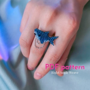 Può includere: Un anello a forma di squalo blu e bianco con una catena d'argento. L'anello è su una mano, e lo sfondo è un tessuto blu chiaro. Modello PDF Tessitura ad angolo retto.