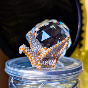 May include: A beaded origami crane with orange and white beads sits on top of a clear glass jar with a silver lid. The crane is perched on top of a clear crystal.