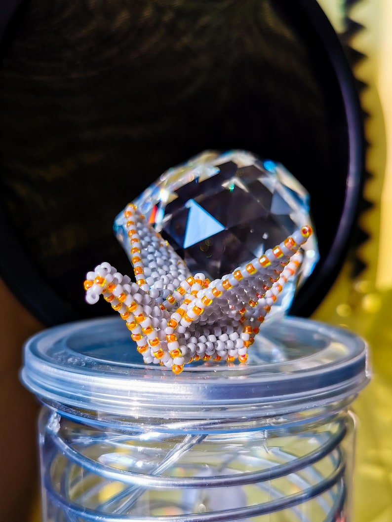 May include: A beaded origami crane with orange and white beads sits on top of a clear glass jar with a silver lid. The crane is perched on top of a clear crystal.