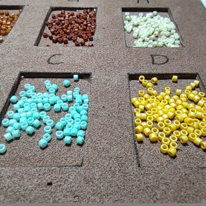 May include: A close-up of ten small containers filled with different colored beads. The containers are labeled with letters A through D. The beads are in various colors, including brown, white, blue, and yellow.