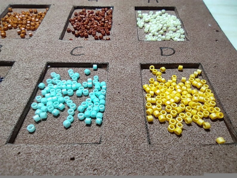 May include: A close-up of ten small containers filled with different colored beads. The containers are labeled with letters A through D. The beads are in various colors, including brown, white, blue, and yellow.