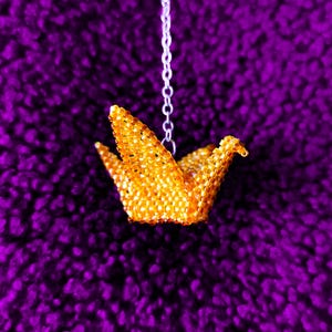 May include: A gold origami crane made of beads hangs from a silver chain against a purple background.