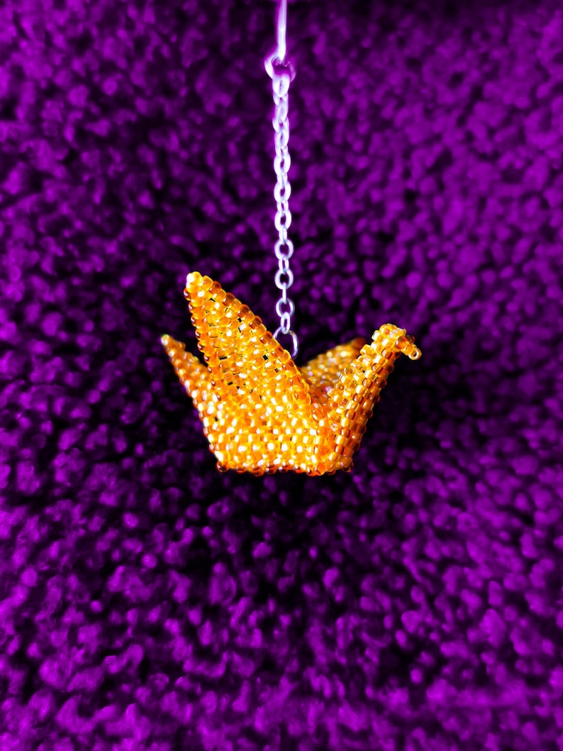 May include: A gold origami crane made of beads hangs from a silver chain against a purple background.