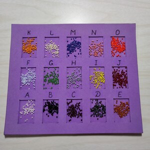 May include: A purple foam board with 15 squares, each containing a different color of small beads. The squares are labeled with letters from A to O.