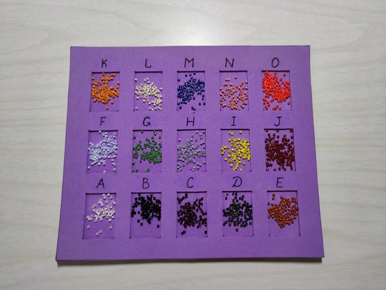 May include: A purple foam board with 15 squares, each containing a different color of small beads. The squares are labeled with letters from A to O.