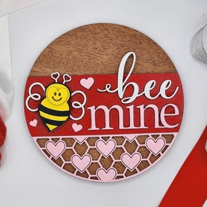 Puede incluir: Un letrero redondo de madera con un patrón de panal rojo y rosa. El letrero dice "bee mine" en letras blancas y presenta una abeja de dibujos animados con una cara amarilla y rayas negras y amarillas.