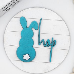 May include: A white wooden round sign with a blue bunny silhouette and the word "hop" in cursive. The bunny has a white fluffy tail.