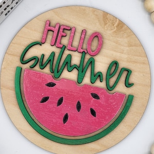 May include: A round wooden sign with the text "Hello Summer" in pink and green letters. A watermelon slice with black seeds is in the centre of the sign.