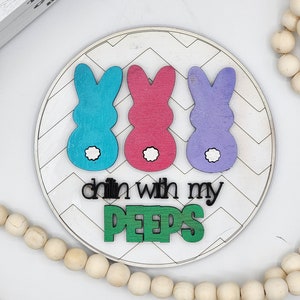 May include: A round wooden sign with a white chevron pattern. Three wooden peep bunnies in blue, pink, and purple are arranged across the top. The text "Chillin with my PEEPS" is written in black and green letters.