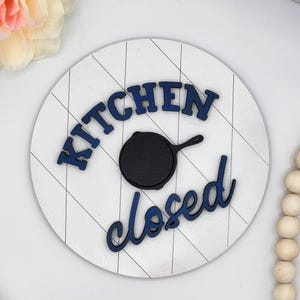 Puede incluir: Un letrero redondo de madera blanca con un patrón de cuadrícula diagonal. Las palabras "KITCHEN" y "closed" están en escritura azul marino. Una sartén de hierro fundido negra está en el centro. Letrero decorativo de cocina.