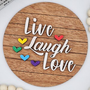 Puede incluir: Un letrero redondo de madera con las palabras "Live Laugh Love" en letras blancas. El letrero tiene un patrón de veta de madera y está decorado con cinco pequeños corazones de varios colores: rojo, amarillo, verde, azul y morado.