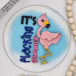 Puede incluir: Un letrero redondo de madera con fondo azul y blanco. El letrero dice "It's a flocking summer" en letras de colores. Un flamenco rosa con pico y patas amarillas está en el letrero.