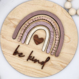 May include: A wooden round sign with a rainbow made of layered wood pieces in shades of pink and brown. The rainbow has a small heart in the center. The words "be kind" are written in cursive below the rainbow.