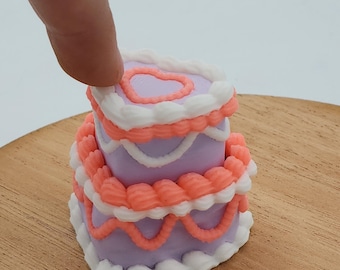 Puede incluir: Una tarta en miniatura de dos pisos en forma de corazón con glaseado lavanda, decorada con glaseado blanco y melocotón. El nivel superior presenta un diseño de corazón. La tarta está sobre una superficie de madera.