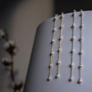 Può includere: Tre delicate collane di perle di diverse lunghezze. Le collane sono esposte su una superficie bianca e presentano una catena dorata con perle bianche e due catene d'argento con perle bianche.