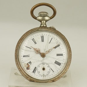 Könnte beinhalten: Antike Taschenuhr mit silberfarbenem Gehäuse und weißem Zifferblatt mit römischen Ziffern und einem Hilfszifferblatt. Die Uhr hat ein dekoratives Design um die Lünette und einen goldfarbenen Aufzugsstift und eine Schlaufe. Das Zifferblatt der Uhr ist gerissen.