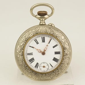 Könnte beinhalten: Silberne Taschenuhr mit weißem Zifferblatt, schwarzen römischen Ziffern und filigranen Zeigern. Die Uhr hat ein verziertes Blumendesign um die Lünette und ein kleines Sekunden-Zifferblatt. Die Aufzugskrone hat eine kontrastierende Bronzefarbe.