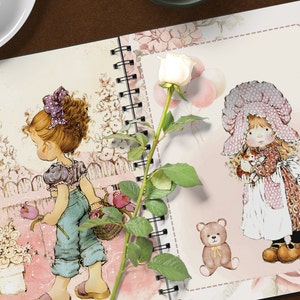 K&ouml;nnte beinhalten: Ein rosa-wei&szlig;es Notizbuch mit floralem Design und zwei Illustrationen von M&auml;dchen. Ein M&auml;dchen h&auml;lt einen Blumenkorb und das andere ein kleines K&auml;tzchen. Eine wei&szlig;e Rose mit gr&uuml;nen Bl&auml;ttern befindet sich auf der linken Seite des Notizbuchs.