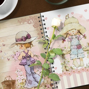 K&ouml;nnte beinhalten: Zwei spiralgebundene Notizb&uuml;cher mit einem rosa-wei&szlig;en Blumendesign. Ein Notizbuch zeigt ein M&auml;dchen mit einem lila Kleid und einem braunen Hut, das Blumen h&auml;lt. Das andere Notizbuch zeigt ein M&auml;dchen mit einem blauen Kleid und einem Strohhut, das auf einem Zaun sitzt. Eine wei&szlig;e Rose mit gr&uuml;nen Bl&auml;ttern liegt zwischen den Notizb&uuml;chern.