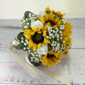 Pode incluir: Um buqu&ecirc; de flores com girass&oacute;is amarelo-brilhantes com centros castanho-escuros, intercalados com pequenas flores brancas de gips&oacute;fila e rosas brancas. O arranjo inclui folhas verdes de eucalipto e uma longa fita na cor creme.