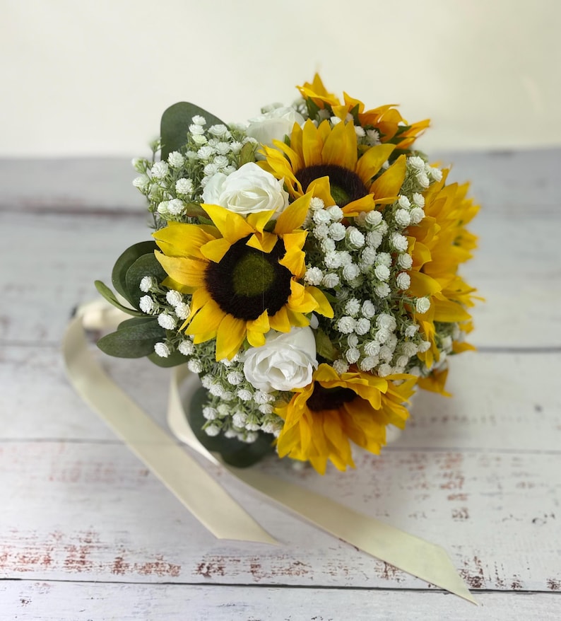 Pode incluir: Um buqu&ecirc; de flores com girass&oacute;is amarelo-brilhantes com centros castanho-escuros, intercalados com pequenas flores brancas de gips&oacute;fila e rosas brancas. O arranjo inclui folhas verdes de eucalipto e uma longa fita na cor creme.