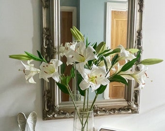 May include: A silver framed mirror with an ornate design hangs above a wooden dresser. A vase with white lilies sits on the dresser, and a wooden sign reads "Our Happy Place".