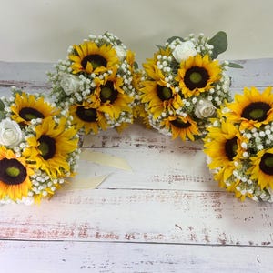 Pode incluir: Tr&ecirc;s ramos de flores com girass&oacute;is amarelo-brilhante, pequenas rosas brancas e delicadas flores de gips&oacute;fila brancas. Cada arranjo inclui folhas de eucalipto verdes e est&aacute; atado com uma fita de cor creme.