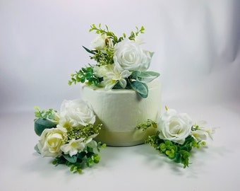 May include: White wedding cake topper featuring three white roses and greenery. The roses are arranged in a cluster on top of the cake, with greenery surrounding them.