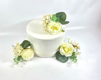 May include: A white cake dummy decorated with three white roses and greenery. The roses are made of fabric and have a realistic appearance.