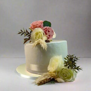Pode incluir: Um bolo branco com um efeito degradê azul claro, decorado com uma fita branca e um buquê de rosas rosa e brancas e ervas secas.
