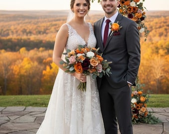 May include: A person in a white lace wedding dress with a v-neckline and a long train, standing next to a person in a charcoal gray suit with a burgundy tie and a boutonniere. They hold a bouquet of orange, burgundy, and white flowers.
