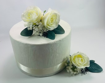 May include: A white cake with a white satin ribbon and a floral topper made of two white roses and greenery.