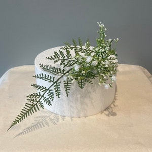 May include: A white round cake dummy with a green fern and white flowers on top.