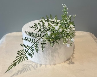 May include: A white round cake dummy with a green fern and white flowers on top.