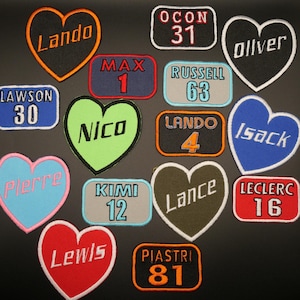 May include: A collection of heart-shaped and rectangular patches with names and numbers. The patches are in various colours, including red, blue, green, black, and white. The names include Lando, Lawson, Pierre, Nico, Isack, Oliver, Lewis, and Lance. The numbers include 30, 31, 1, 63, 4, 12, 16, and 81.