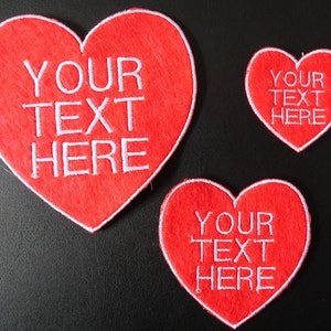 May include: Three red felt heart-shaped patches with white embroidered text. The largest patch reads "YOUR TEXT HERE". The other two patches are smaller and read "YOUR TEXT HERE".