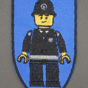 May include: Embroidered patch depicting a Lego-style police officer in a black uniform and helmet, on a blue shield. The patch has a black border and is suitable for application on clothing or accessories. The patch measures approximately 7 cm in height.